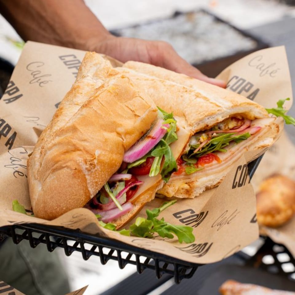 A hand holds a Cafe COPA baguette sandwich filled with meat, cheese, red onion, and arugula.