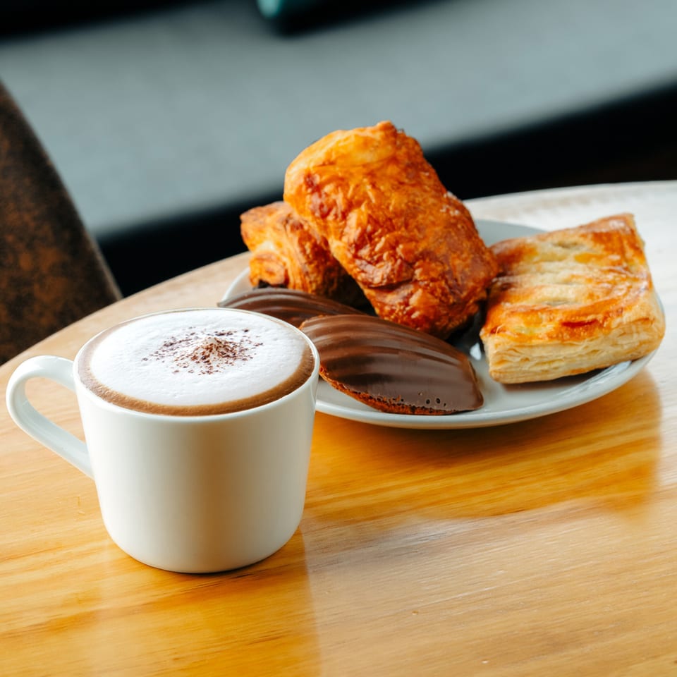 A Cafe COPA cappuccino with foam and cocoa powder, next to pastries and chocolate madeleines.