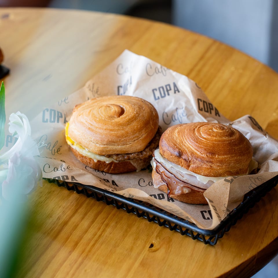 Two Cafe COPA croissant sandwiches with egg and cheese in a basket on a wooden table.