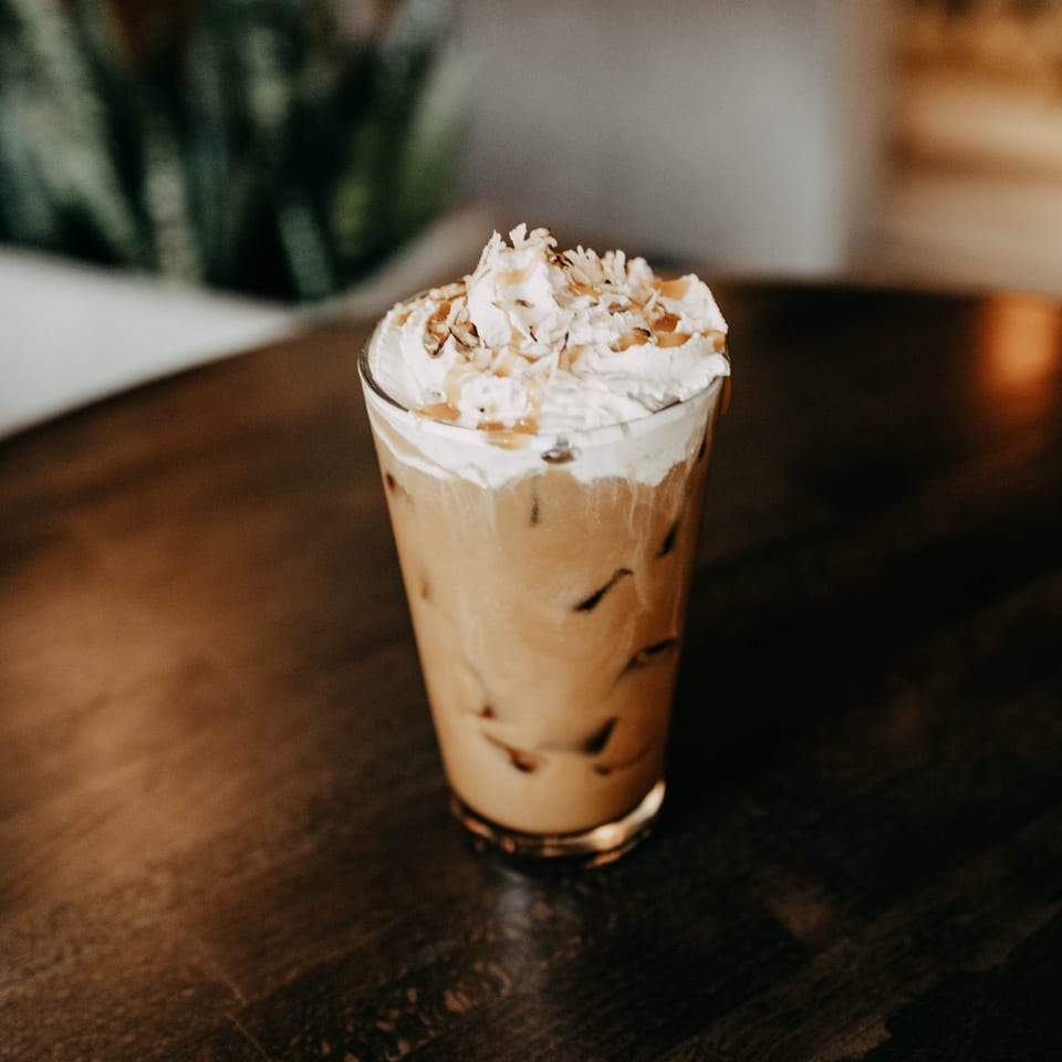Iced coffee with whipped cream, caramel drizzle, and nuts on a dark wooden table.