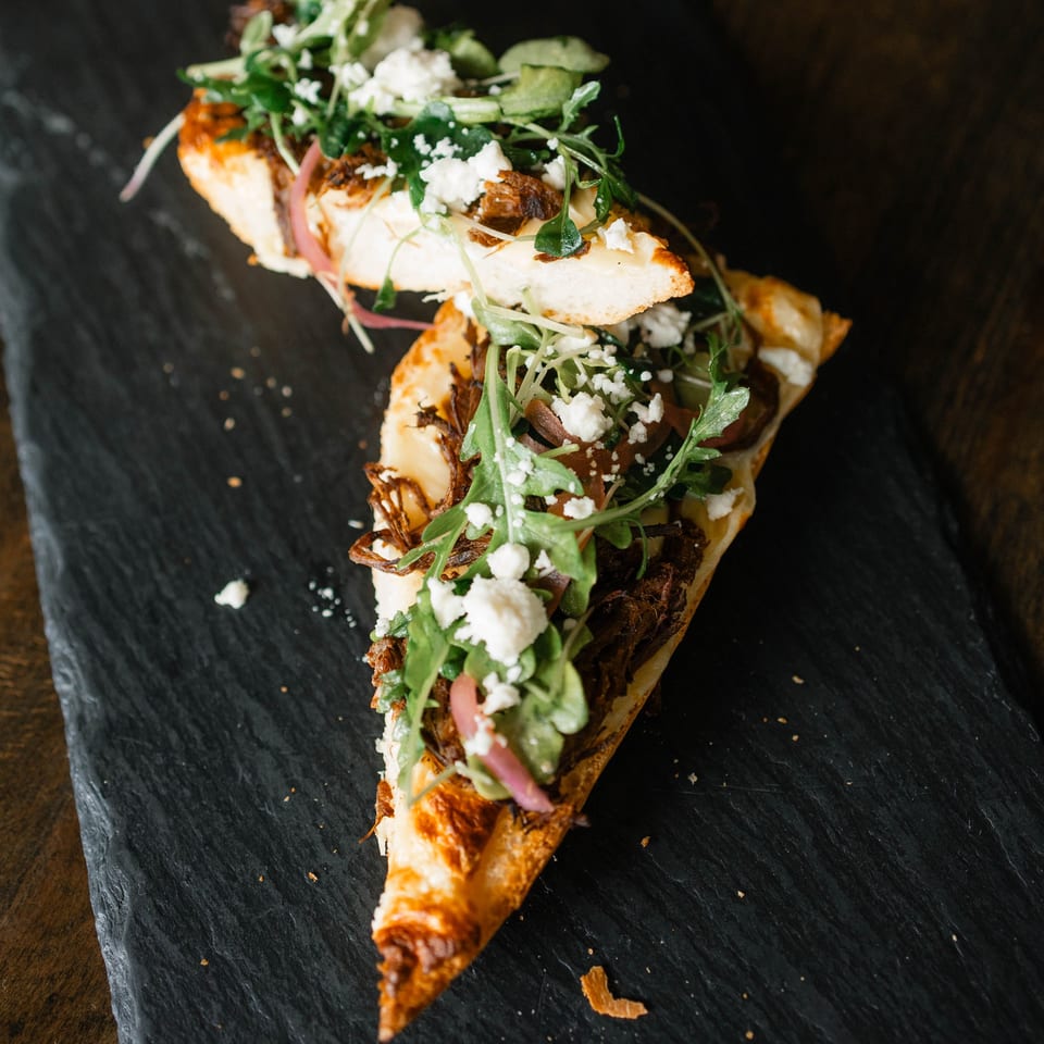 Two slices of flatbread pizza topped with shredded meat, arugula, and feta cheese on a dark slate.