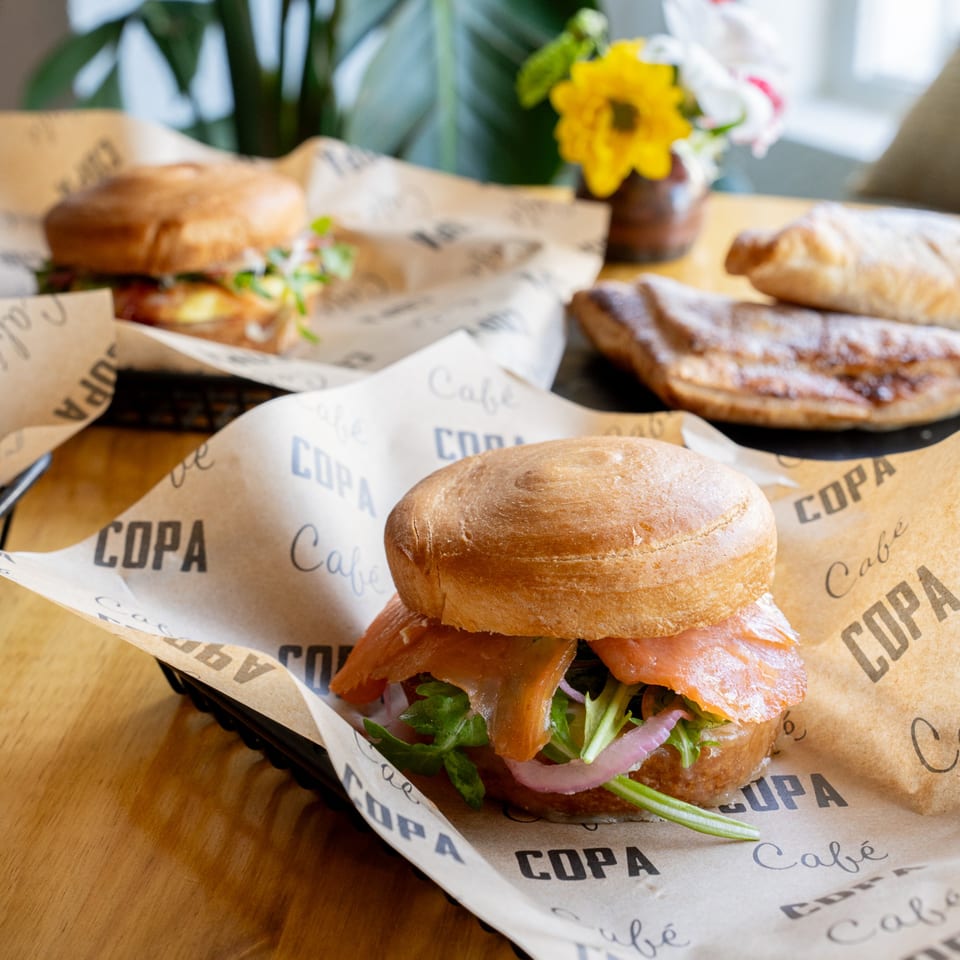 Cafe COPA sandwiches with salmon, arugula, and red onion on branded paper, with pastries in the background.