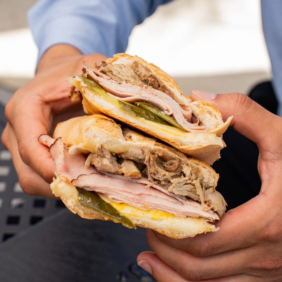 Hands holding a Cafe COPA Cuban sandwich, revealing layers of ham, pork, pickles, and cheese.