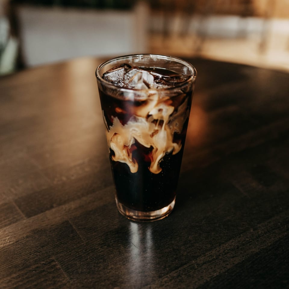 Iced coffee with cream swirling into dark liquid, on a dark wooden table at Cafe COPA.