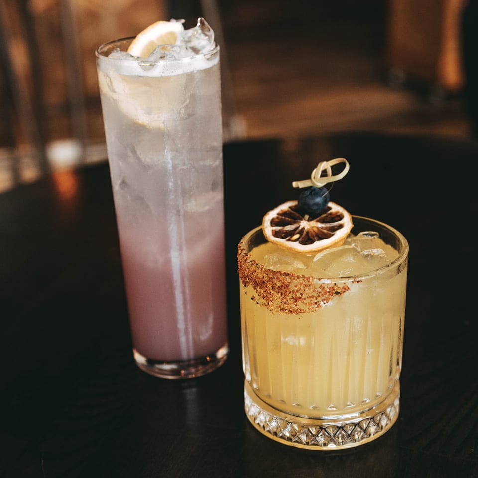 Two Cafe COPA cocktails, one purple with lemon, one yellow with a spiced rim and dried orange.