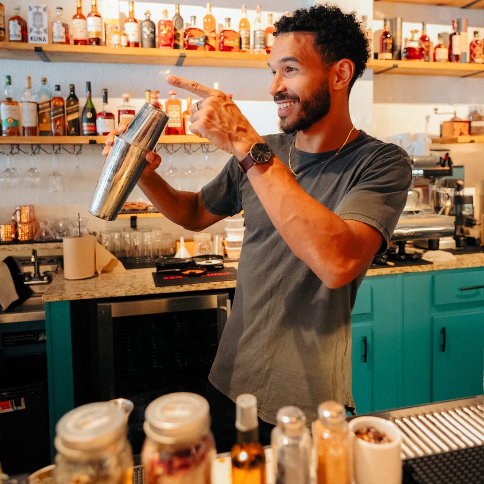 A smiling Cafe COPA bartender shakes a cocktail shaker behind a bar with liquor bottles.